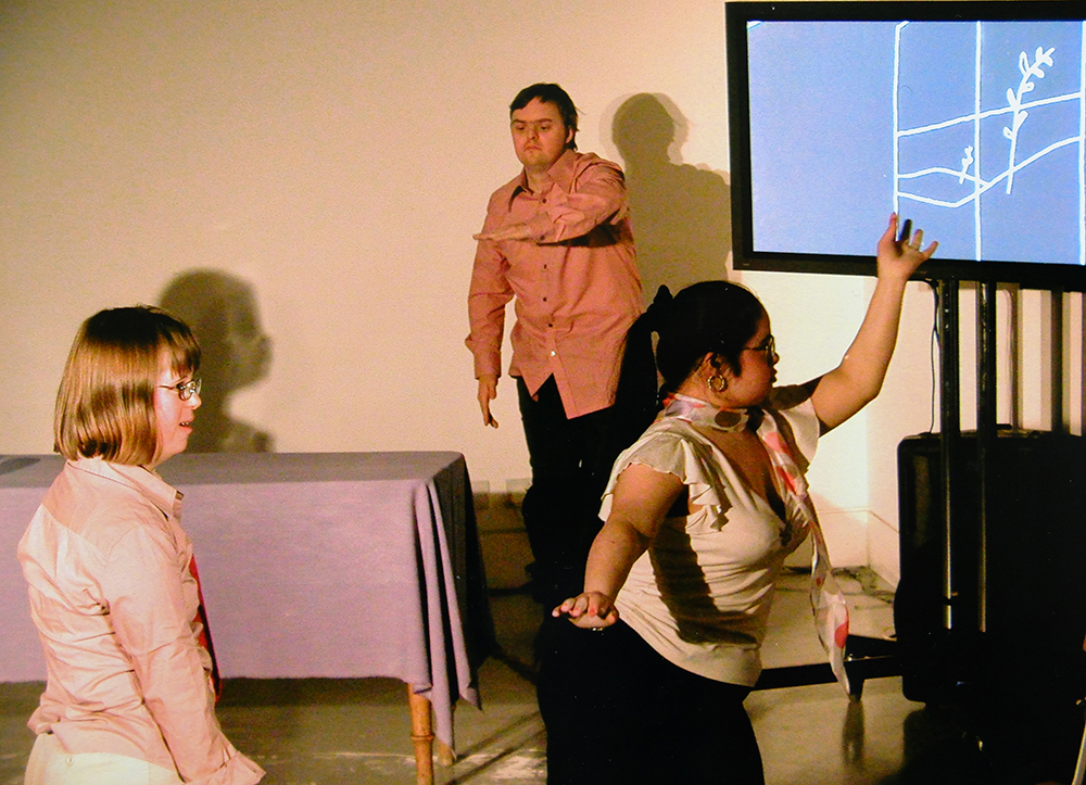 A still image from the performance Kissed by Corali Dance Company at the main gallery space of Café Gallery Projects, featuring Bethan Kendrick, John Long and Aisha Booth. Aisha dancers in front of a flat screen monitor which features an image of a plant, she holds her arm up to echo the shape of the plant stem. They wear pink and cream costumes.